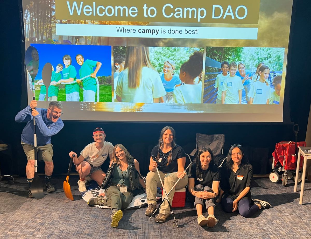 DAO staff, Mike, Elle, Megan, Ally, Marisha, and Shilpa dressed in camping attire posed with camping gear (paddles, innertubes, trekking poles, and camping chairs) in front of a 'Welcome to Camp DAO; where campy is done best' sign.
