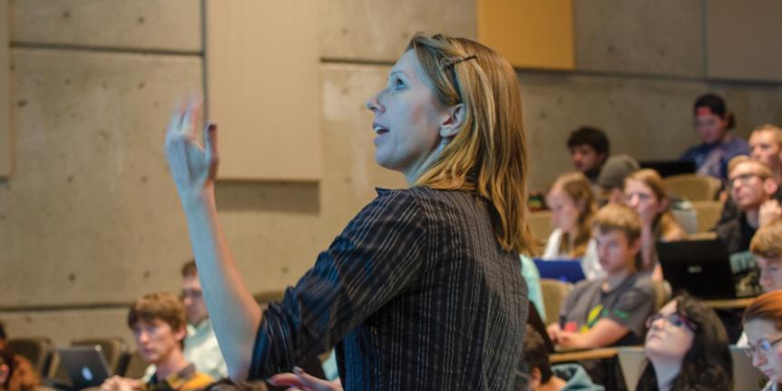 An instructor looks toward a screen while teaching a class in a large lecture hall on the CU Boulder campus.