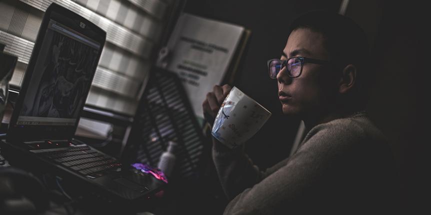 Man drinks coffee and watches a video on a laptop in a darkened room.