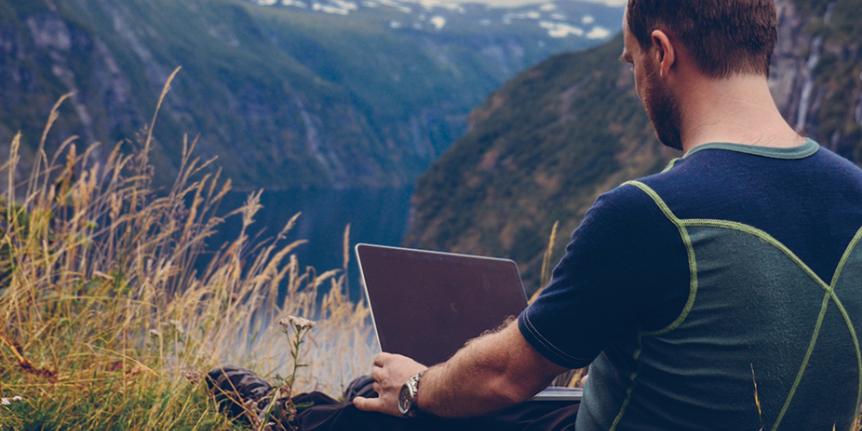 Man sitting with laptop on ridge overlooking a fjord.